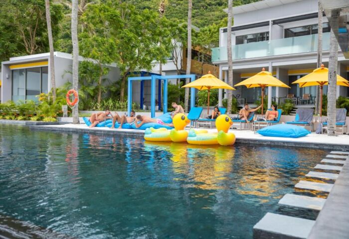 Explorar Koh Phangan adults-only resort pool scene. Gay-friendly beach hotel with men relaxing, yellow umbrellas, and duck floats.