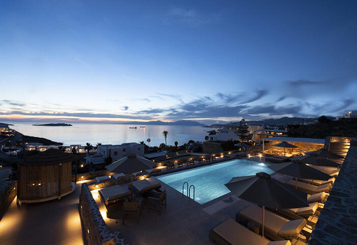 Mykonos Bay Resort & Villas pool view at dusk. Luxury beachfront gay hotel in Mykonos Town. Sunbeds and umbrellas near the pool.