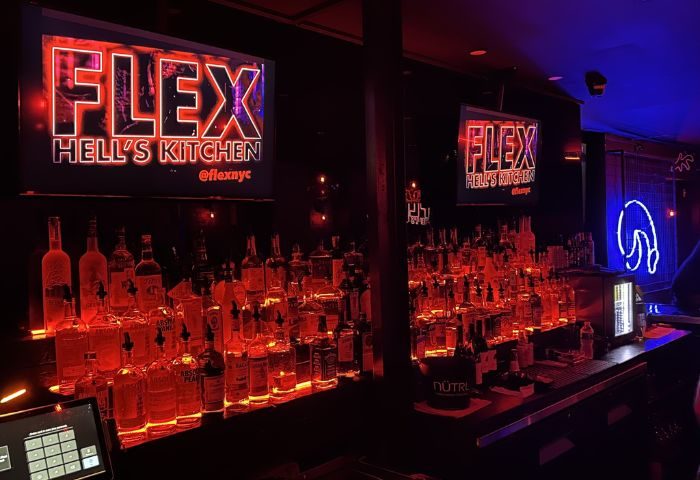 Bar at FLEX Gay Bar in New York. Shelves are full of liquor bottles. Neon signs say FLEX Hell's Kitchen. Red and blue lighting.