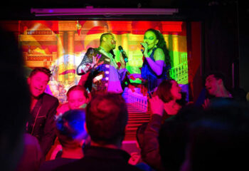 FLM Hong Kong gay bar: Karaoke night with drag queens performing on stage. Crowd enjoys the show in a vibrant, colorful atmosphere.