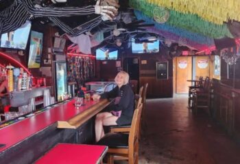 Interior of Falcon bar in Long Beach, a gay-friendly travel destination. A patron sits at the red bar, rainbow decor above.