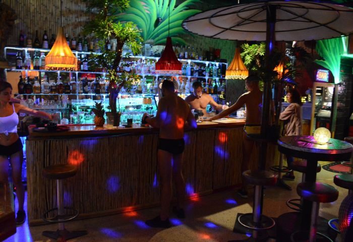 Fiction gay bar in Gran Canaria. Shirtless bartenders serve drinks in a tropical-themed gay bar with colorful lights. Gay nightlife.