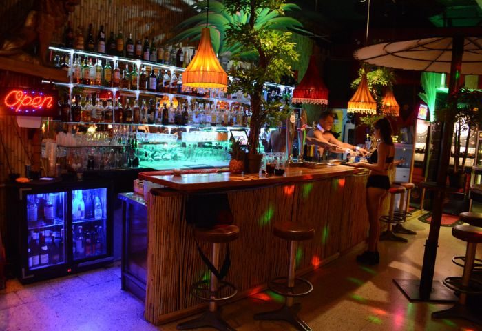 Fiction gay bar in Gran Canaria. Bartender serving drinks at a tiki-style bar with tropical decor, neon lights, and bar stools.