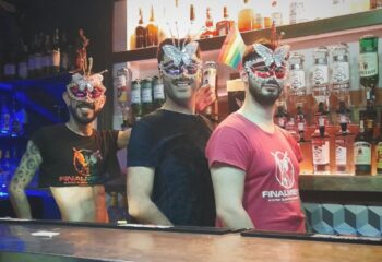 Three gay men in butterfly masks at Finalmente Club, Lisbon. Bartenders smile in front of liquor bottles. Gay travel advice.