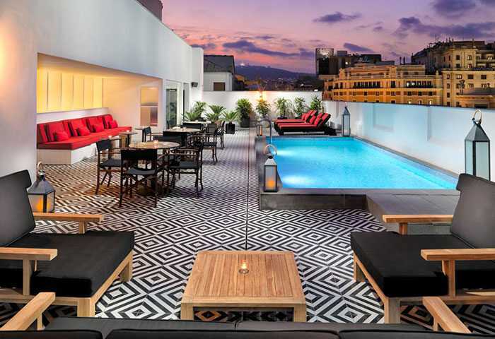 H10 Metropolitan Hotel rooftop pool in Barcelona at dusk. Gay hotel with cheap price, lounge chairs, tables, and city views.