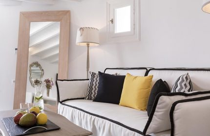 Absolute Mykonos Suites room for 4 people in Mykonos Town Gayborhood. Couch with pillows, fruit and drinks on table, mirror and lamp.