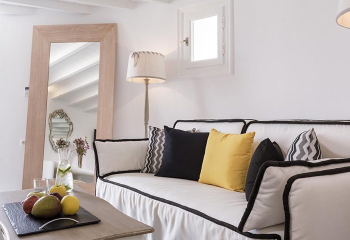 Absolute Mykonos Suites room for 4 people in Mykonos Town Gayborhood. Couch with pillows, fruit and drinks on table, mirror and lamp.