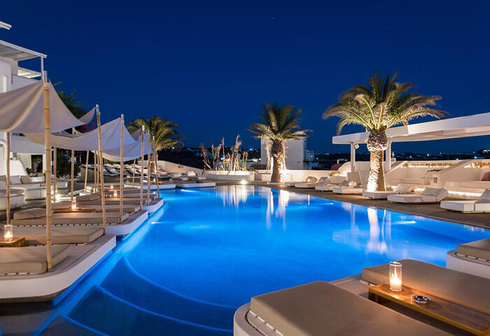 Andronikos Hotel's rooftop pool at night. Luxury gay hotel with last-minute availability. Palm trees, lounge chairs, and blue-lit pool.