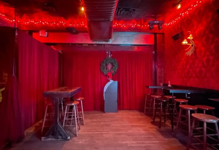 Interior of Flaming Saddles Gay Bar in New York. Red lighting, bar stools, tables, and stage with red curtains and a wreath. Gay travel advice.