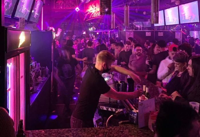 Flash on Church, a gay bar in Toronto. Bartender serving drinks in a crowded, lively space with purple lighting and disco balls.