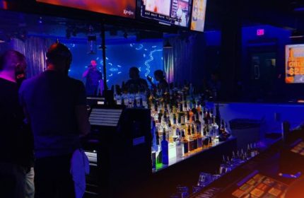Flex Cocktail Lounge in Las Vegas. Interior view of the bar area with bottles, screens, and people in a dimly lit, blue-toned space.