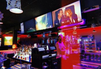 Flex Cocktail Lounge in Las Vegas. Bartender at the bar, multiple screens showing movies. Red and purple lighting. Gay bar travel.