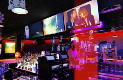 Flex Cocktail Lounge in Las Vegas. Bartender at the bar, multiple screens showing movies. Red and purple lighting. Gay bar travel.