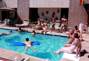 Flex LA: Gay men relax by a pool. Some lounge on rafts, others sit poolside at this gay travel destination. Rainbow flag visible.
