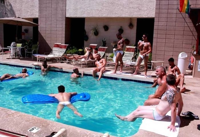 Flex LA: Gay men relax by a pool. Some lounge on rafts, others sit poolside at this gay travel destination. Rainbow flag visible.