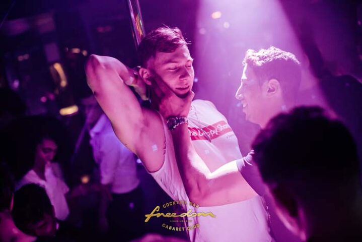 Freedom Bar Soho London: Two men interact at the gay bar, one near a pole, in a vibrant, nightlife setting. Gay travel advice.