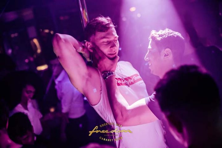 Freedom Bar Soho London: Two men interact at the gay bar, one near a pole, in a vibrant, nightlife setting. Gay travel advice.