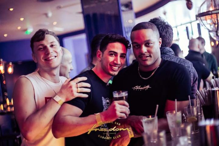 Three smiling men pose at Freedom Bar Soho, a gay bar in London. Cocktails and bar equipment visible. Gay travel.