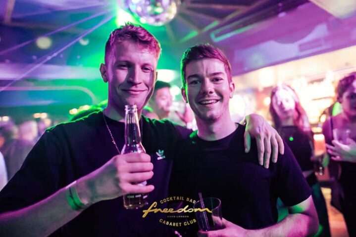 Two gay men smiling at Freedom Bar Soho, London. Nightlife scene with disco ball and purple lighting. Gay travel destination.
