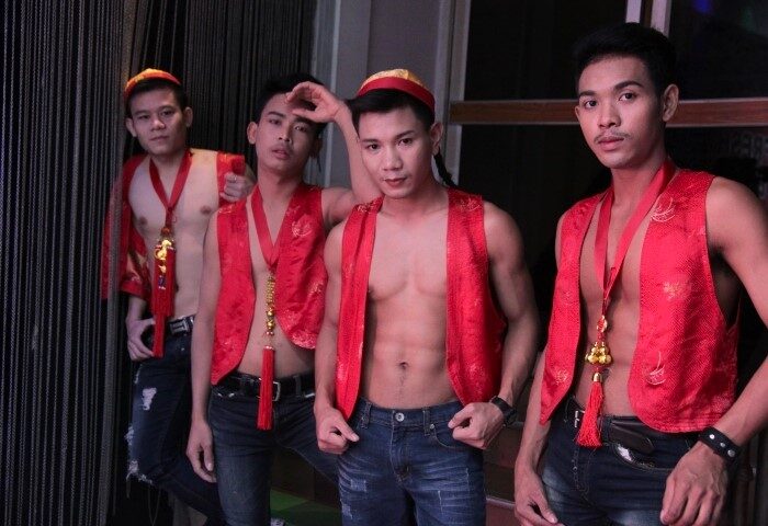 Bangkok Go-Go Boys: Four fresh-faced, shirtless men in red vests and hats, posing at a gay bar. Nightlife in Thailand for gay travelers.