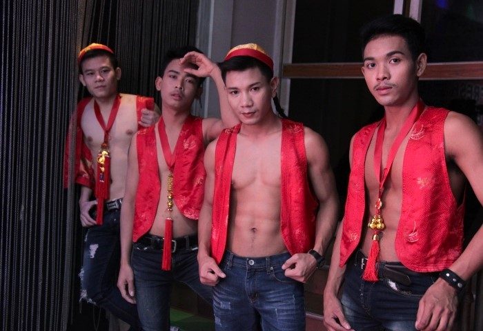 Bangkok Go-Go Boys: Four fresh-faced, shirtless men in red vests and hats, posing at a gay bar. Nightlife in Thailand for gay travelers.