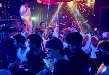 Frolic Bar Ho Chi Minh: A crowded gay bar scene with men dancing under neon lights. A performer stands on the bar. Gay travel advice.