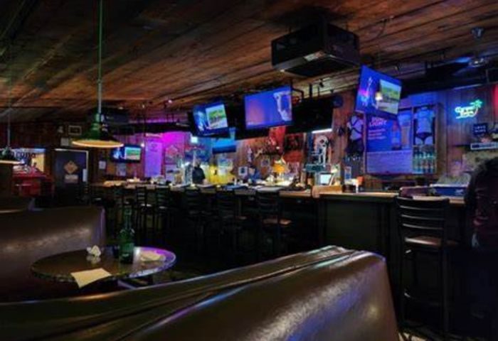 Interior of Fun Hog Ranch, a gay bar in Las Vegas. TVs, bar stools, and a leather booth are visible. The bar has a rustic, inviting vibe.