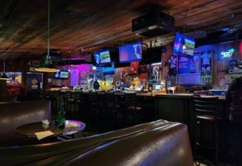 Interior of Fun Hog Ranch, a gay bar in Las Vegas. TVs, bar stools, and a leather booth are visible. The bar has a rustic, inviting vibe.