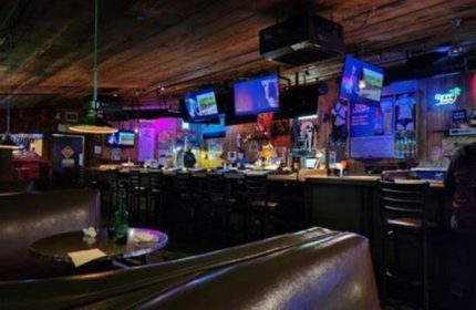 Interior of Fun Hog Ranch, a gay bar in Las Vegas. TVs, bar stools, and a leather booth are visible. The bar has a rustic, inviting vibe.