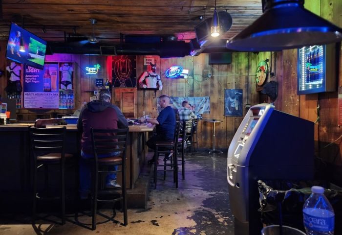 Interior view of Fun Hog Ranch gay bar in Las Vegas. Patrons at the bar, neon signs, and rustic wood paneling. Gay travel guide.