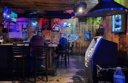 Interior view of Fun Hog Ranch gay bar in Las Vegas. Patrons at the bar, neon signs, and rustic wood paneling. Gay travel guide.