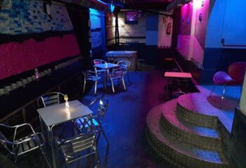 Interior of GRIS Gay Bar in Madrid. Tables, chairs, and a small stage are illuminated with blue and pink lighting. Gay travel advice.