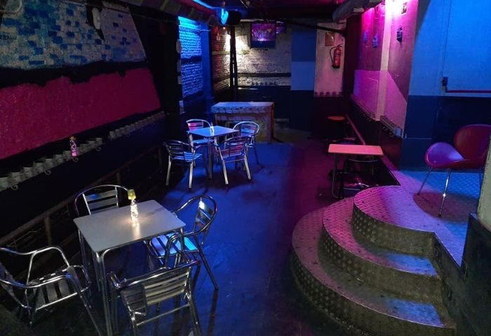Interior of GRIS Gay Bar in Madrid. Tables, chairs, and a small stage are illuminated with blue and pink lighting. Gay travel advice.
