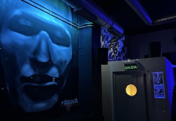 Interior of GRIS Gay Bar in Madrid. Blue-lit wall mural of a face, exit door with Salida sign. Gay travel advice.
