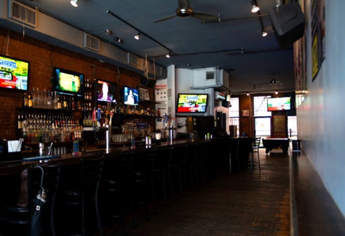NYC gay sports bar interior. Long bar with stools, TVs showing sports, liquor shelves, and a pool table in the back. Gay travel advice.