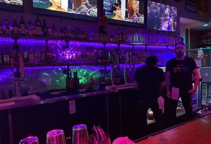 GYM Sportsbar: Bar with purple lights, bottles, & TVs. Bartender wearing a GYM shirt. Popular gay sports bar for gay men travelers.