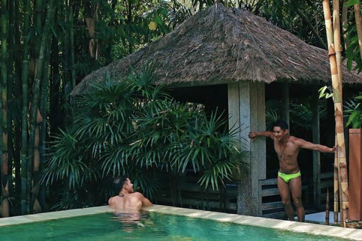Gay Bali Ubud hotel pool scene. Two men, one in the pool, other by a thatched hut. Lush greenery, tropical vibes, gay travel.