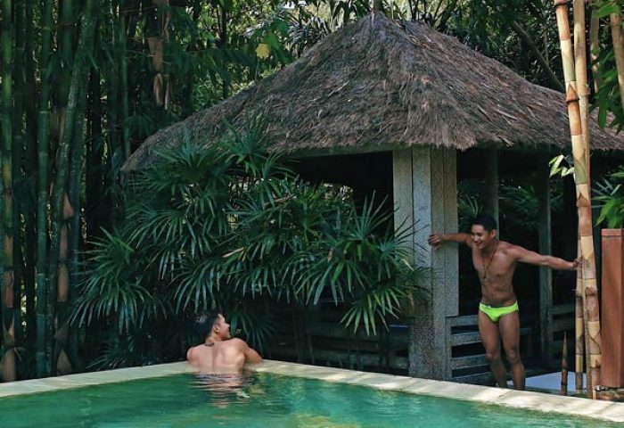 Gay Bali Ubud hotel pool scene. Two men, one in the pool, other by a thatched hut. Lush greenery, tropical vibes, gay travel.