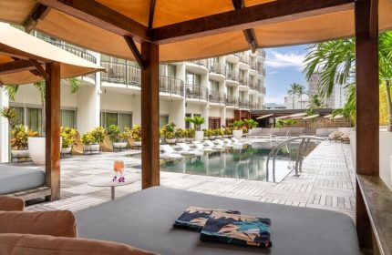 The Laylow Autograph Collection gay-friendly hotel pool view. Cabana with lounge chairs, towels, and drinks. Balconies and palm trees visible.