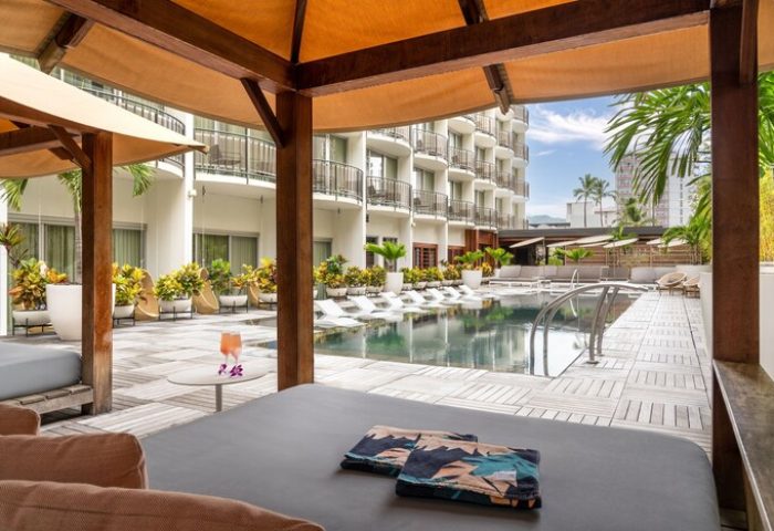 The Laylow Autograph Collection gay-friendly hotel pool view. Cabana with lounge chairs, towels, and drinks. Balconies and palm trees visible.
