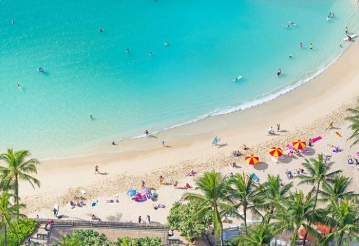 Aerial view of Waikiki Beach Tower, a gay-friendly hotel. Turquoise water, sandy beach, palm trees, and people swimming and sunbathing.