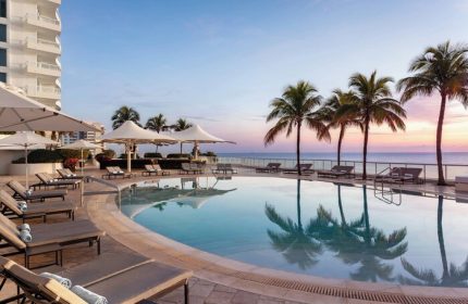 The Ritz-Carlton, Fort Lauderdale gay-friendly hotel pool view. Palm trees, lounge chairs, and ocean at sunset. A luxury travel destination.