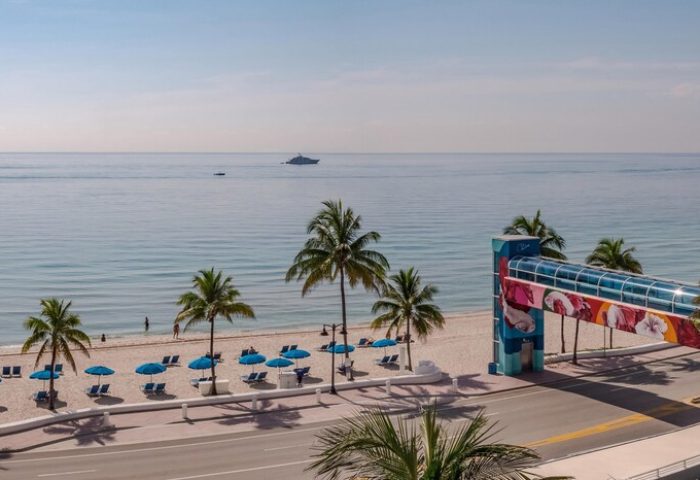 The Westin Fort Lauderdale Beach Resort view. Gay-friendly hotel with beach access via walkway. Palm trees, ocean, and blue umbrellas.