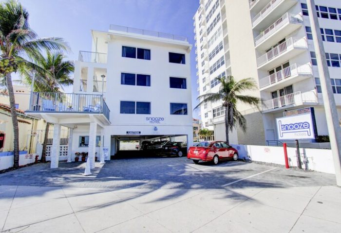 Snooze Hotel, a gay-friendly hotel. White building with parking, palm trees, and a sign. A red car is parked outside. Great for gay travel.