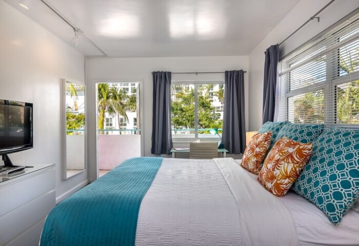 Nobleton Hotel gay-friendly room. Bright, airy bedroom with balcony. Palm trees visible. Comfortable bed with decorative pillows. Modern, clean design.