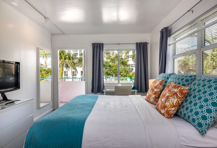 Nobleton Hotel gay-friendly room. Bright, airy bedroom with balcony. Palm trees visible. Comfortable bed with decorative pillows. Modern, clean design.