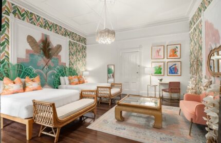 Marrero's Guest Mansion gay-friendly hotel room. Two beds, rattan benches, colorful art, leaf wallpaper, and a peach chair create a stylish space.