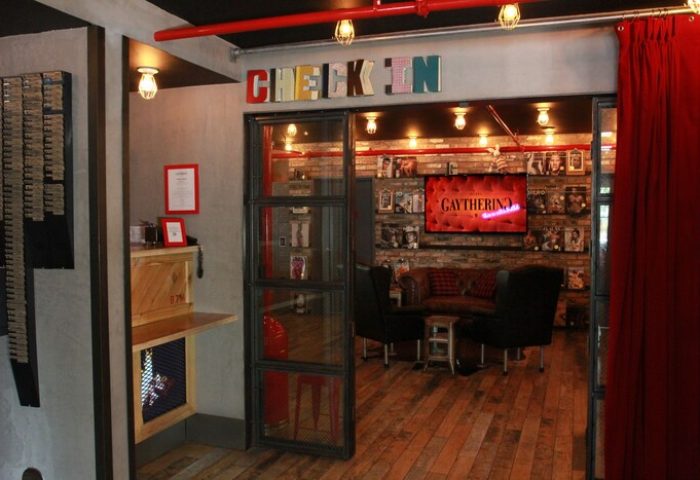 The Gaythering, a gay-friendly hotel: check-in area with a TV screen displaying Gaythering. Crates and vintage decor add to the ambiance.