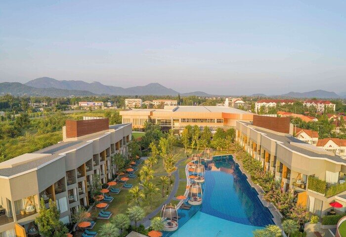 Avani+ Hua Hin Resort aerial view. Gay-friendly hotel with pool, lounge chairs, and mountain backdrop. Thailand travel destination.