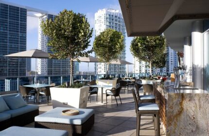 Kimpton EPIC Hotel's rooftop bar. A gay-friendly hotel with city views, tables, chairs, umbrellas, trees, and a bar area. Miami travel.